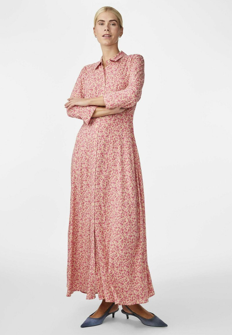 Robe maxi florale rose avec un design à boutons, des manches longues et une silhouette fluide. Portée avec des talons aiguilles bleus.