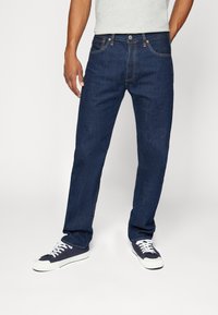 Hombre con jeans rectos de color azul oscuro y zapatillas azul marino con suelas y cordones blancos, de pie contra un fondo neutro.