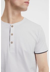White short-sleeve henley shirt with three brown buttons and a black accent at the sleeve hems. Smooth cotton fabric, crew neckline.