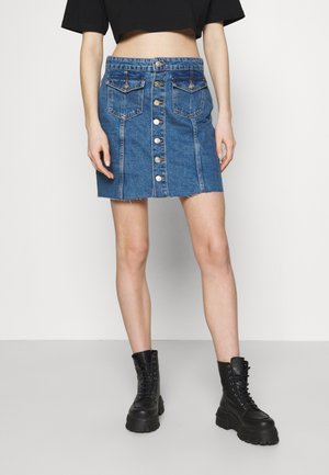 Kvinde iført en blå denimnederdel med knapper foran og forlommer, sort crop top og sorte snørebånds støvler, stående foran en hvid baggrund.