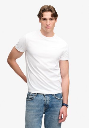Giovane uomo con capelli castani che indossa una semplice maglietta bianca a maniche corte, jeans blu e un orologio da polso blu, che posa con una mano dietro la schiena.