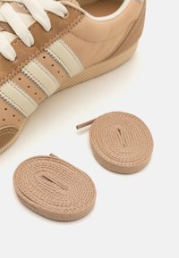 Brązowe trampki z białymi paskami i perforowanymi detalami. Dwie skręcone brązowe sznurówki na pierwszym planie, teksturowane wzorem plecionki.