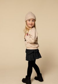 Meisje met blond haar, gekleed in een beige gebreide muts, beige sweatshirt, zwarte rok, zwarte panty en zwarte laarzen, staande in profiel op een beige achtergrond.