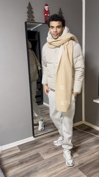 Lichtbeige donsjack met een hoge kraag, crèmekleurige sjaal, grijze joggingbroek en witte sportschoenen. Spiegelreflectie zichtbaar op de achtergrond.