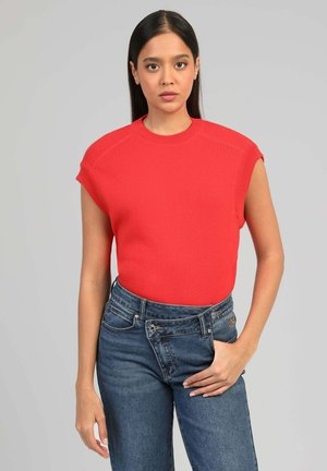 Femme aux longs cheveux noirs portant un haut rouge sans manches et un jean bleu, debout avec une main près de la poche, fond neutre.