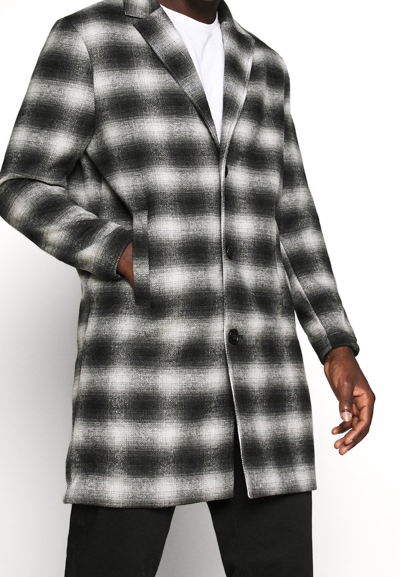 Cappotto a quadri bianco e nero con taglio dritto, caratterizzato da doppi bottoni, rever a punta e tasche laterali. Realizzato in tessuto strutturato.