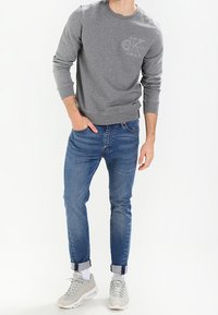 Sudadera gris con cuello redondo, mangas con puños y un pequeño logo en el pecho, combinada con jeans ajustados azules y zapatillas claras.