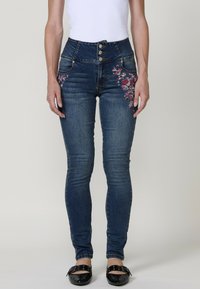 Vaqueros de tiro alto en azul con bordado floral en la pierna derecha, presentan cierre de botón y un ajuste slim, confeccionados en denim.