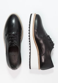 Black leather shoes with a rounded toe, lace-up design, and a white, textured sole. Features a contrasting tan accent around the sole.