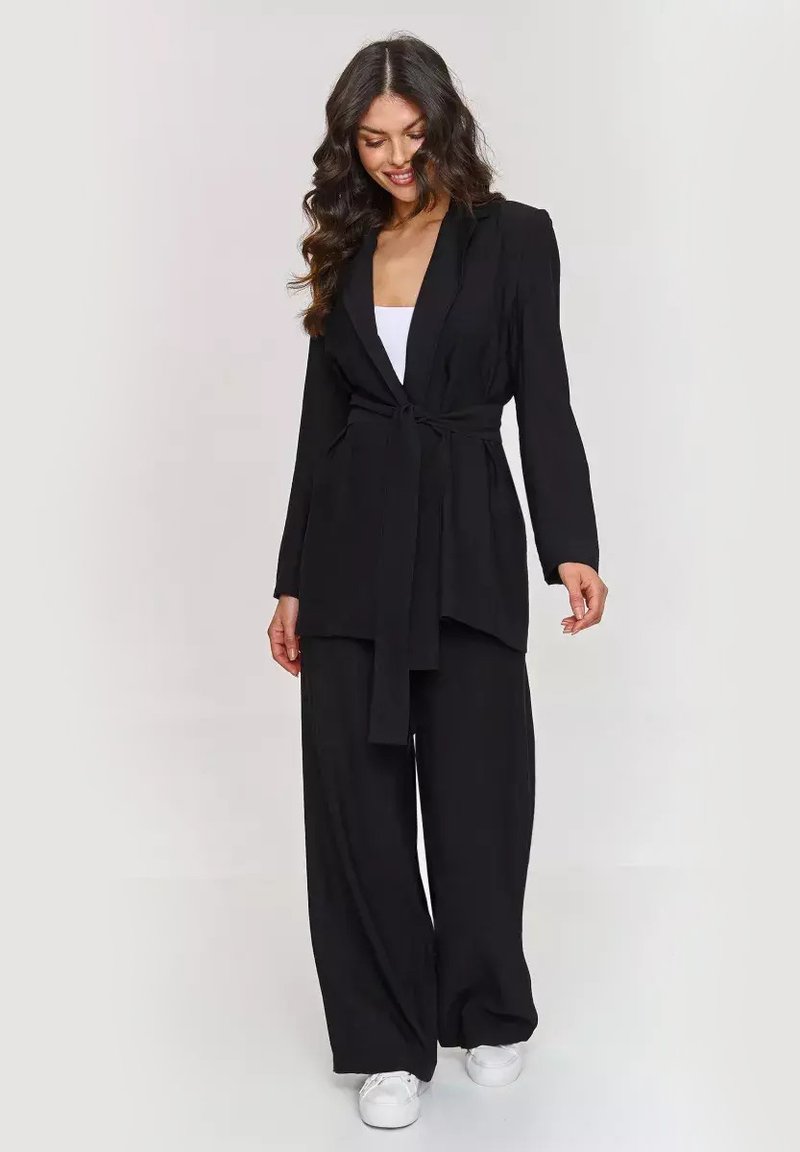 Czarna marynarka z wiązaniem w talii, długimi rękawami i dopasowanym krojem, zestawiona z szerokimi czarnymi spodniami i białymi butami. Gładka struktura tkaniny.