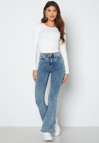 Vit ribbad långärmad crop top som kombineras med högmidjade blå bootcut-jeans. Modellen bär vita sneakers; enkla silverörhängen som accenter.