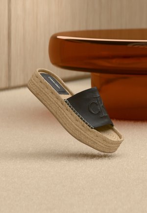 Beige platformsandal med vævet sål og sort overdel i læder prydet med "CK"-logo, placeret nær et brunt rundt bord.