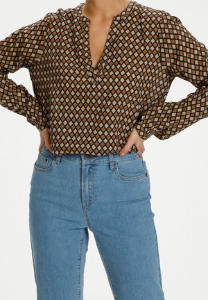 Femme portant un chemisier marron à manches longues avec un motif en losanges, rentré dans un jean taille haute bleu clair, les mains posées sur les hanches.