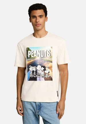 Junger Mann trägt ein weißes T-Shirt mit buntem Peanuts-Cartoon-Motiv, das eine Straße überquert, und blaue Jeans.