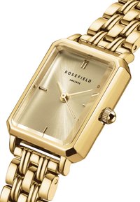 Reloj rectangular dorado con una esfera de oro cepillado, que presenta marcadores finos y una corona; conectado por una pulsera de metal dorado pulido.