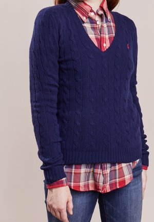 Personne portant un pull en maille torsadée bleu marine à col en V par-dessus une chemise à carreaux rouge et bleu boutonnée, avec un jean bleu, debout devant un fond uni.