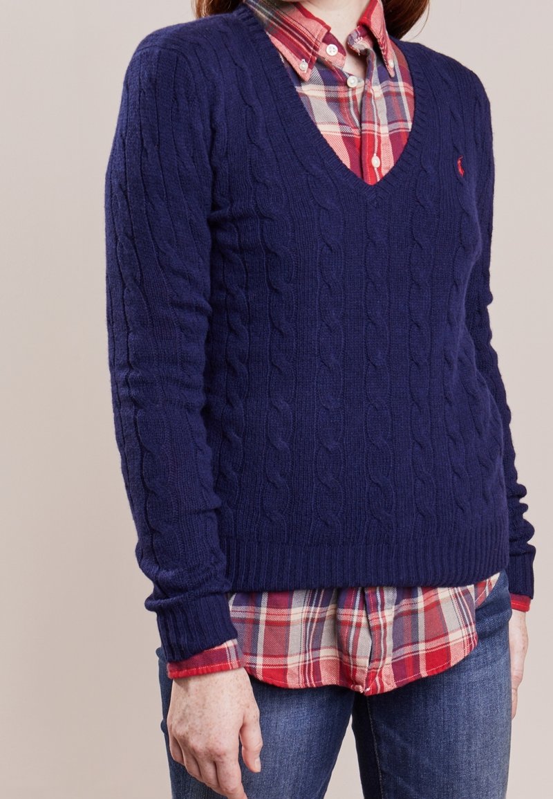 Personne portant un pull en maille torsadée bleu marine à col en V par-dessus une chemise à carreaux rouge et bleu boutonnée, avec un jean bleu, debout devant un fond uni.