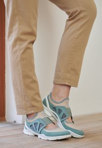 Lightweight athletic shoe featuring a breathable mesh upper in teal and beige with adjustable strap, contrasting white sole, and padded collar.