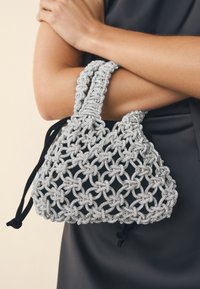 Sidabrinė rankinė su karoliukais, nėriniuota raštu, faktūruotu paviršiumi ir dviem pintais rankenomis. Uždarymas su raišteliais, juodos spalvos akcentais.