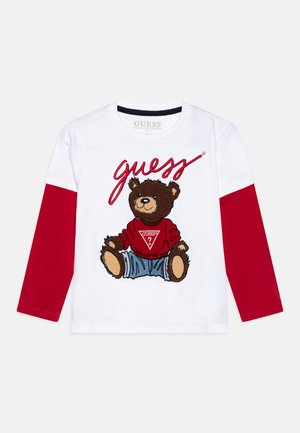 Weißes Langarmhemd mit einem braunen Teddybär-Motiv, roten Ärmeln und dem Schriftzug "guess" in einer geschwungenen Schriftart. Hergestellt aus Bio-Baumwolle.