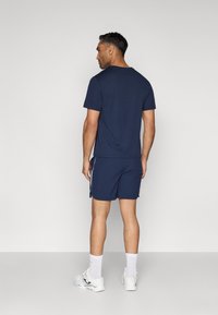 Tamsiai mėlynas marškinėlis su apvaliu kaklu, derinamas su atitinkančiomis šortais, kurių šonai dekoruoti baltais raštais. Modelis dėvi baltus sportinius batus ir kojines.