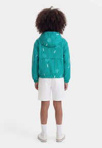 Παιδί που στέκεται με την πλάτη, φορώντας πετρόλ μπουφάν με κουκούλα και κατακόρυφες λευκές ρίγες και σχέδιο με ρακέτα του τένις, λευκά σορτς, κάλτσες και αθλητικά παπούτσια.