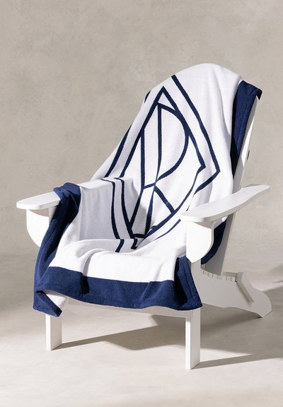 Λευκή και ναυτική κουβέρτα με γεωμετρικό μοτίβο κρεμασμένη πάνω σε μια λευκή ξύλινη καρέκλα. Η υφή φαίνεται απαλή και αφράτη, προσφέροντας μια ζεστή εμφάνιση.