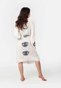 Biela bavlnená róba so zelenými a čiernymi motívmi očí, dlhými rukávmi a strapcami na spodnom leme. Navrhnuté pre pohodlie a ľahké nosenie.
