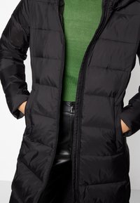 Czarna puchowa kurtka z zamkiem błyskawicznym, długimi rękawami i bocznymi kieszeniami. Nosi się ją na zielonym dzianinowym topie o gładkiej, błyszczącej fakturze.