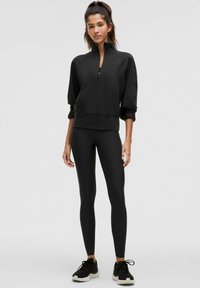 Top deportivo negro con cremallera y cuello alto, combinado con leggings negros y zapatillas negras. Material suave, diseño ajustado y acabado sin costuras.