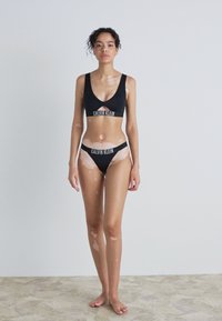 Melna sporta krūšturis un bikini komplekts ar izgriezumiem, ar logotipa joslu. Gluda auduma, minimālisma dizains un kontrastējošas ādas krāsas.