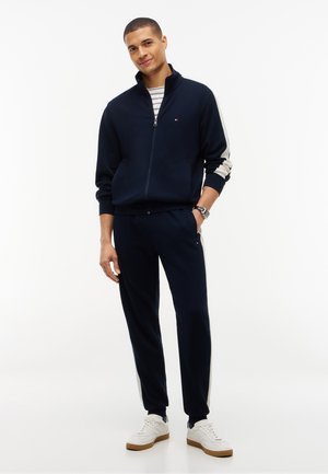 Jeune homme debout, portant un survêtement bleu marine avec des bandes blanches sur les manches et des baskets blanches, souriant légèrement avec une main dans la poche.