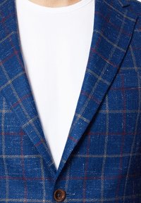 Niebieska marynarka w kratę wykonana z teksturowanej tkaniny z brązowym guzikiem. Wewnętrzna warstwa zawiera białą koszulę. Wyraźny wzór w kratkę w kolorach czerwonym i beżowym.