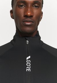 Czarna kurtka zapinana na zamek, wykonana z gładkiego, elastycznego materiału. Posiada wysoki kołnierz oraz biały detal z logo na froncie.