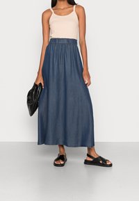 Denim-maksihame, jossa on joustava vyötärö, yhdistettynä beigeen toppiin. Mustat ristikkäin kulkevat sandaalit ja teksturoitu musta clutch-laukku.