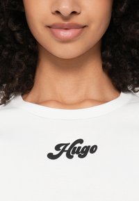 Balta kokvilnas T-kreklu ar melnu "Hugo" logo, kas ir izliekta dizainā. Apaļš izgriezums un gluda tekstūra, piemērota ikdienas valkāšanai.