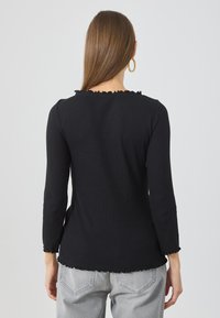 Black ribbed long-sleeve top with ruffled neckline and hem, paired with light gray jeans. Smooth texture and fitted design.