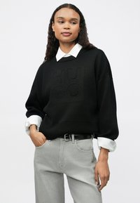 Maglione nero testurizzato con logo in rilievo, camicia bianca con colletto sotto e jeans grigio chiaro, con cintura nera e vestibilità rilassata.