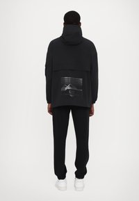 Svart huva jacka med en reflekterande ryggtryck som visar berg, matchad med svarta sweatpants och vita sneakers. Slät textur.
