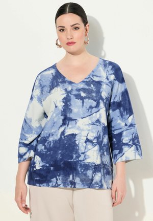 Svetr s modrým tie-dye vzorem, výstřihem do V a rukávy tří čtvrtin. Obsahuje texturované akcenty a jemné dírkové vzory po stranách.