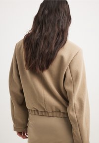 Mujer con cabello largo y ondulado de color oscuro viste una chaqueta corta beige sobre una falda beige a juego, de pie frente a un fondo claro y liso.
