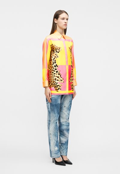 Γυναίκα όρθια, φορώντας μπλούζα με μακριά μανίκια που έχει πάνελ σε κίτρινο, ροζ και λεοπάρ μοτίβο, σε συνδυασμό με ξεθωριασμένο μπλε τζιν και μαύρες μυτερές γόβες.