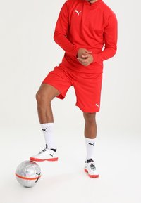 Person in roter Sportkleidung kontrolliert einen silbernen und orangefarbenen Fußball mit weißen Fußballschuhen vor einem schlichten weißen Hintergrund.