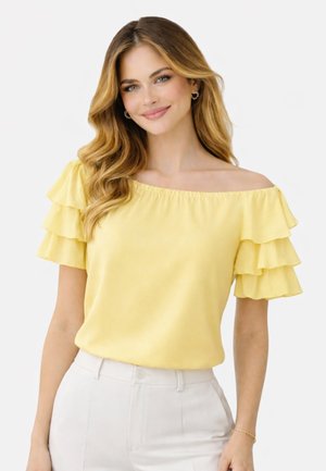 Młoda kobieta o blond, falowanych włosach ubrana w żółtą bluzkę z opuszczonymi ramionami i falbankami na rękawach oraz białe spodnie, uśmiechająca się do aparatu.