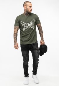 Olive zelené bavlnené tričko s veľkým bielym logom "TAPOUT", skombinované s čiernymi roztrhnutými džínmi a bielymi teniskami. V ruke má čiernu čiapku.