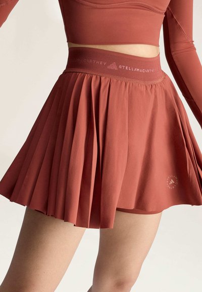 Frau trägt einen rostfarbenen, plissierten Sportrock mit integriertem Shorts und ein passendes langärmliges Crop-Top, steht vor einem einfarbigen Hintergrund.