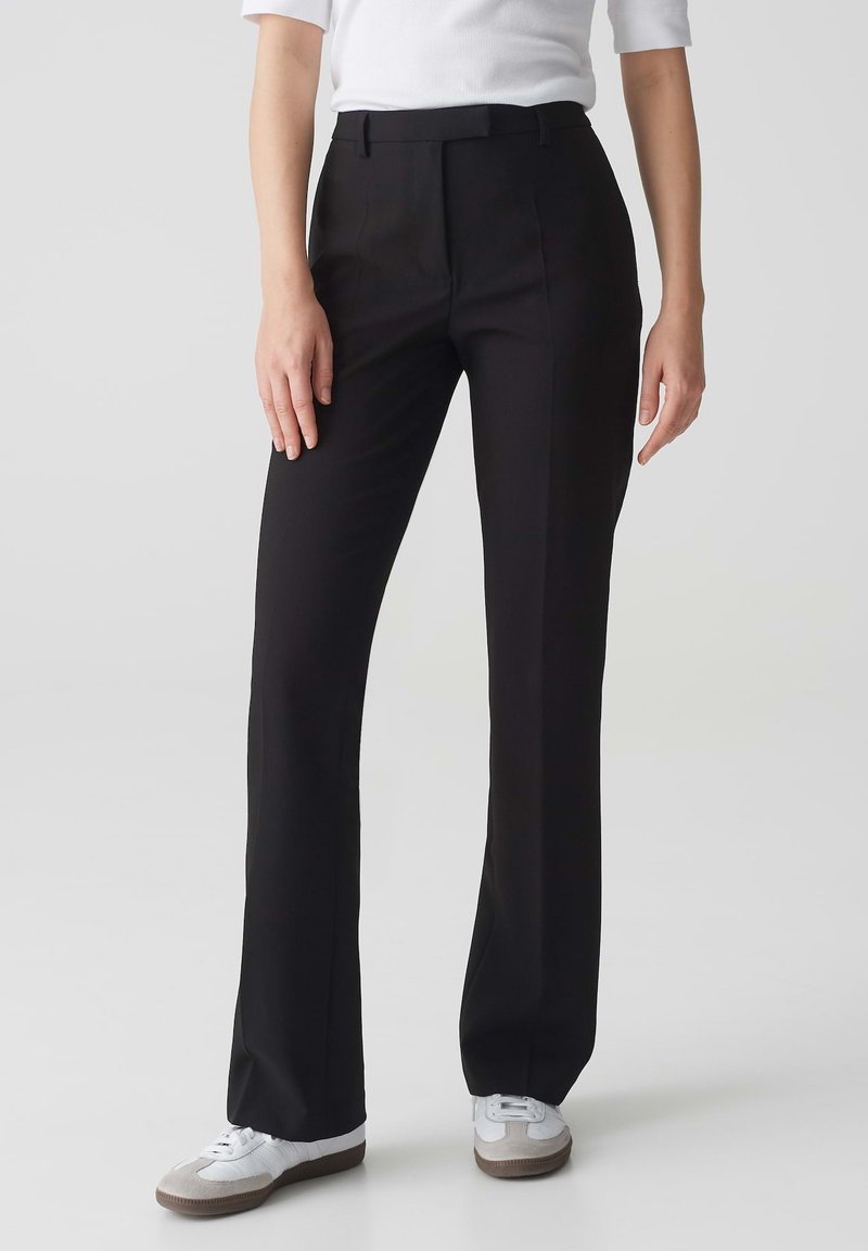Pantalon noir avec une coupe ajustée, devant plat et passants de ceinture, associé à des baskets blanches ; tissu lisse avec un léger brillant.
