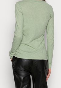 Langärmliges T-Shirt in hellgrün mit rundem Halsausschnitt, aus weichem Stoff gefertigt. Kombiniert mit hochgeschnittenen schwarzen Lederhosen mit Gesäßtaschen.