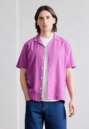 Νεαρό άτομο με μεσαίου μήκους σκούρα μαλλιά, που φοράει ένα ανοιχτό μωβ κοντομάνικο πουκάμισο πάνω από ένα λευκό μπλουζάκι και μπλε τζιν, όρθιο.