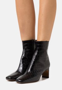 Botas negras hasta el tobillo hechas de cuero con estampado de cocodrilo, con puntera afilada y tacón cuadrado. Detalle de cremallera en el lado.